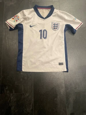 England fotbollströja Bellingham #10 - Säljer en vit England fotbollströja från Nike med blå detaljer och nummer 10 på bröstet och ryggen. Bellingham står tryckt på ryggen. Tröjan har officiella landslagsmärket på bröstet och EM-märke på ärmen. Perfekt för dig som älskar fotboll och landslaget.