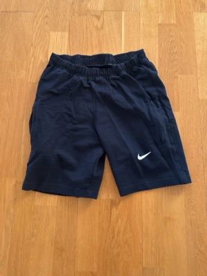 Marinblå shorts från Nike - Säljer ett par marinblå shorts från Nike med elastisk midja och den klassiska vita Nike-loggan på benet. Shortsen har en avslappnad passform och är perfekta för träning eller chill. Tillverkade i mjukt bomullsmaterial.