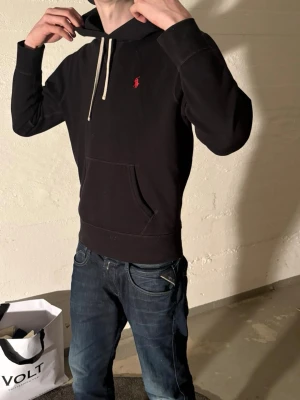 Svart hoodie från Polo Ralph Lauren - Svart hoodie från Polo Ralph Lauren med klassisk röd logga broderad på bröstet. Hoodien har vit dragsko i huvan och en stor magficka framtill. Tillverkad i mjukt bomullsmaterial och har ribbade muddar vid ärmslut och nederkant.modellen på bilden är 187 cm och väger ca 70 kilo. Hoodien är köpt på NK i stockholm, kvitto finns vid begär. 