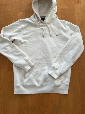 Vit hoodie från Gant med logga - Snygg vit hoodie från Gant med liten broderad logga på bröstet. Klassisk modell med huva och dragsnören samt magficka framtill. Tillverkad i mjukt bomullsmaterial och har ribbade muddar vid ärmar och nederkant. Perfekt för en avslappnad och stilren look.