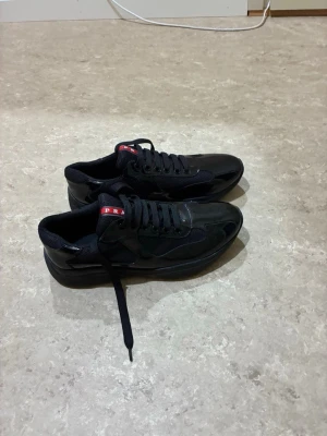 Svarta sneakers från Prada - Snygga svarta sneakers från Prada med blank finish och meshdetaljer. Skorna har klassisk låg profil, svarta skosnören och en röd logga på plösen. Perfekta för dig som vill ha en stilren och exklusiv look.helt oanvänd. 