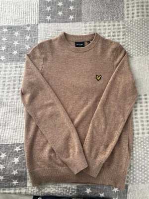Beige stickad tröja från Lyle & Scott - Stickad tröja i beige från Lyle & Scott med rund halsringning och lång ärm. Tröjan har den klassiska gula örnloggan broderad på bröstet och ribbade muddar vid ärmslut och nederkant. Perfekt för lager på lager och enkel att matcha.