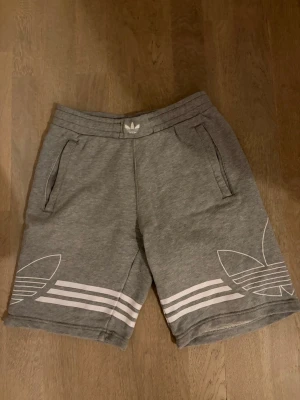 Grå Adidas shorts med vita detaljer - Snygga grå shorts från Adidas med vita ränder och stora loggor på benen. Mjukt material och bekväm passform med resår i midjan och två fickor. Perfekta för en avslappnad stil.