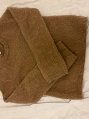 Beige fluffig stickad tröja H&M - Mysig beige stickad mohair tröja från H&M Edition i fluffigt material. Tröjan har rund halsringning, långa ärmar och en avslappnad passform. Perfekt för lager på lager och ger en trendig look till hösten och vintern. 