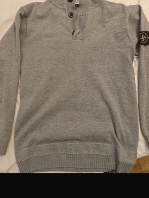 Grå stickad tröja Stone Island - Snygg grå stickad tröja Stone Island med ribbstickade muddar och nederkant. Tröjan har hög krage med knappslå och två knappar framtill samt den klassiska Stone Island-loggan på ärmen. Perfekt för dig som gillar stilrena plagg.