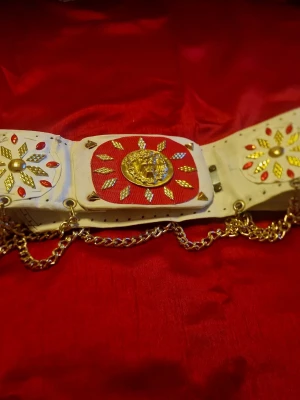 Vitt Elvisbälte med gulddetaljer och kedjor - Unikt vitt bälte med dekorativa gulddetaljer, röda inslag och kedjor. Bältet har ett stort guldfärgat lejonmotiv framtill och mönstrade plattor med röda och guldiga dekorationer. Perfekt för dig som vill sticka ut med accessoarer.