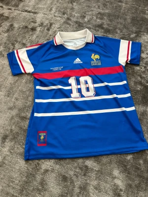 Frankrike Zidane VM 98 fotbollströja - Klassisk blå fotbollströja från Adidas, Frankrikes landslag, VM 1998. Nummer 10 och Zidane på ryggen, vita och röda detaljer, broderad tupp-logga och FIFA World Cup-märke. Tillverkad i lätt och ventilerande material, perfekt för fotboll eller samling.