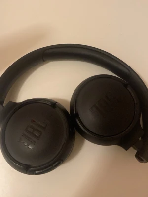 Svarta over-ear hörlurar från JBL - Snygga svarta over-ear hörlurar från JBL med vadderade kåpor och justerbart huvudband. Hörlurarna har en stilren design i plast och konstläder, med tydliga L och R-markeringar på insidan. Perfekta för musik, gaming eller poddar.