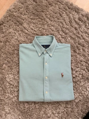 Ljusblå skjorta från Ralph Lauren - Klassisk ljusblå skjorta från Ralph Lauren med button-down krage och broderad logga på bröstet. Skjortan har vita knappar framtill och är tillverkad i mjuk bomull. Perfekt för en clean och stilren look.