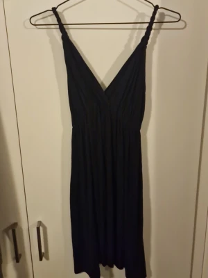 Svart kort klänning med v-ringning - Snygg svart kort klänning med djup v-ringning och breda axelband. Klänningen har en luftig passform och är perfekt för varma dagar. Materialet känns mjukt och följsamt, vilket gör den bekväm att bära. Enkel men stilren design som passar till många tillfällen.