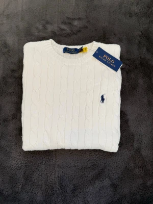 Vit kabelstickad tröja Polo Ralph Lauren - Kabelstickad vit tröja från Polo Ralph Lauren med klassisk rund halsringning och marinblå broderad logga på bröstet. Tröjan har ribbade muddar och är tillverkad i mjukt material som ger en clean och tidlös look.