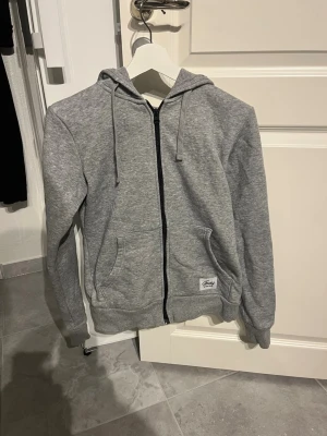 Grå zip hoodie med huva - Snygg gråmelerad hoodie med dragkedja från Fresh. Klassisk modell med två fickor fram, justerbar huva med snören och ribbade muddar. Perfekt för en chill och avslappnad stil. Tillverkad i mjukt bomullsmaterial.