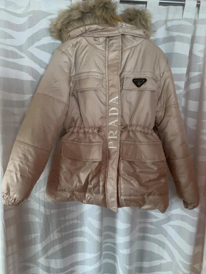 Beige jacka med fuskpäls  - Snygg beige jacka med löstagbar huva och fuskpälsdetalj. Jackan har dragsko i midjan, två bröstfickor med lock och två stora fickor framtill. klassisk trekantslogga på bröstet. 