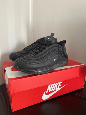 Nike Air Max 97 Triple Black sneakers - Nike Air Max 97 Triple Black sneakers med ikoniska vågiga paneler, helsvart ovandel i mesh och syntet, samt synlig Air-sula. Liten vit Swoosh på sidan och tungan. Skön dämpning och sportig look som passar till streetwear.