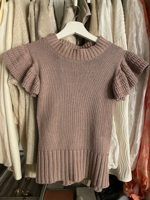 Stickad topp med volangärm Holly & Whyte - Supersöt stickad topp i dovt lila från Holly & Whyte by Lindex. Modellen har korta volangärmar, ribbad halsringning och bred ribbad kant nedtill. Perfekt till jeans eller kjol för en trendig look.