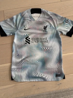 Liverpool Nike bortatröja mönstrad - Säljer en Liverpool FC bortatröja från Nike med ett unikt, psykedeliskt mönster i vitt, svart, blått och lila. Tröjan har svarta muddar, klubbmärke och sponsortryck på bröstet. Perfekt för dig som vill sticka ut på planen eller läktaren.