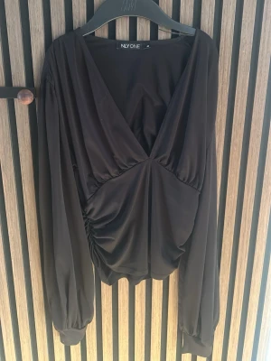 Svart omlottopp från NLY One - Trendig svart omlottopp från NLY One med djup v-ringning och långa puffärmar. Toppen har snygg rynkning i midjan och är tillverkad i ett mjukt, följsamt material som ger en elegant siluett. Perfekt för att lyfta vilken outfit som helst.