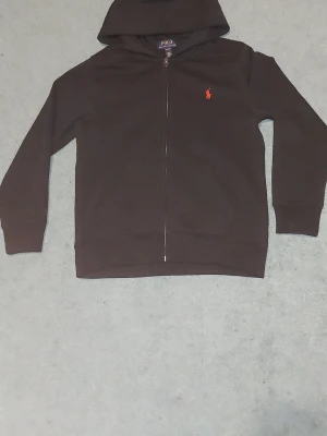 Svart hoodie från Polo Ralph Lauren - Klassisk svart hoodie från Polo Ralph Lauren med dragkedja framtill och huva. Tröjan har det ikoniska röda Polo-märket broderat på bröstet och två fickor fram. Tillverkad i mjukt bomullsmaterial som känns skönt mot huden. Perfekt för en avslappnad och stilren look.