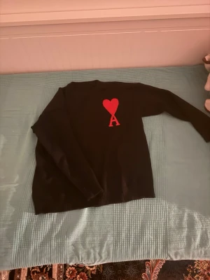 Svart stickad tröja Ami Paris - Svart stickad tröja från Ami Paris med röd hjärtlogga och bokstaven A på bröstet. Klassisk rund halsringning och långa ärmar. Perfekt för dig som gillar stilrena plagg med en twist.