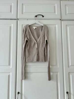 Beige ribbad kofta med v-ringning💖 - Trendig beige ribbad kofta med djup v-ringning och knäppning framtill. Långärmad modell i mjukt, stretchigt material som sitter snyggt på kroppen. Perfekt att styla över ett linne eller t-shirt.