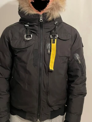 Svart Parajumpers pufferjacka med päls - Säljer en storlek Medium svart pufferjacka från Parajumpers med gul detaljrem, stor huva med beige fuskpäls och flera fickor. Jackan har patch på ärmen, dragkedja framtill och robusta metalldetaljer. Perfekt för kalla vinterdagar.