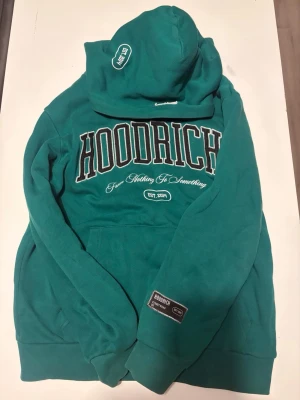 Grön hoodie från Hoodrich - Snygg grön hoodie från Hoodrich med stor logga och text på bröstet samt tryck på ryggen. Klassisk känguruficka framtill, ribbade muddar och huva med tryck. Perfekt för dig som gillar streetwear och vill sticka ut med en bold look. Unisex men mest man