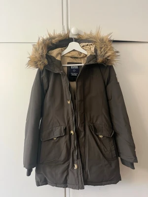 Mörkgrön parkas från Svea med fuskpäls - Stilren mörkgrön parkas från Svea med avtagbar huva och fuskpäls. Jackan har stora fickor framtill, dragkedja och knappar, samt Svea-logga på ärmen. Perfekt för kalla dagar och riktigt mysig att bära.