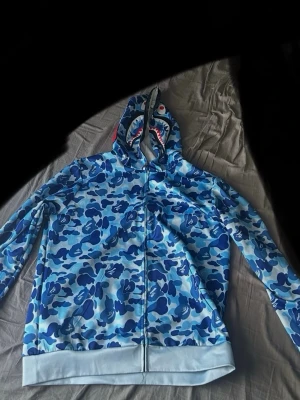 Blå bape hoodie - Säljer en riktigt fet blå camo zip hoodie från BAPE med hajmotiv på huvan. Hoodien har dragkedja hela vägen, ribbade muddar och det klassiska BAPE-mönstret i olika blå nyanser. Perfekt för dig som vill sticka ut med streetwear-vibe.