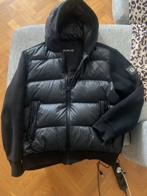 Svart pufferjacka från Bvaldi - Snygg svart pufferjacka/cardigan från Bvaldi med quiltad front och huva. Ärmarna är stickade och har ribbade muddar, medan kroppen är vadderad för extra värme. Två dragkedjeförsedda fickor fram och en patch på ärmen ger en cool detalj.