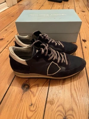 Svarta sneakers från Philippe Model - Säljer ett par svarta Philippe Model sneakers med grå detaljer och vit sula. Skorna har en mix av mocka och mesh, samt den klassiska sköldloggan på sidan. Låga i modellen och med grå snören. Perfekta för dig som gillar stilrena och sportiga sneakers. Endas skor kommer med