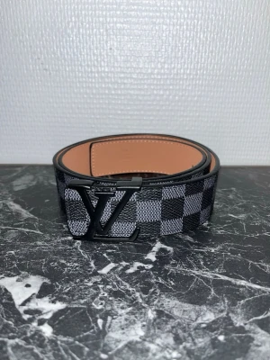 Rutmönstrat bälte i svart och grå - Snyggt bälte med klassiskt rutmönster i svart och grå. Bältet har en stor logga i svart metall som spänne och insidan är i beige skinn. Perfekt accessoar för att lyfta din outfit.