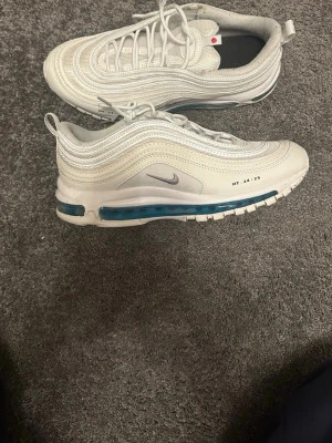 Nike Air Max 97 vita sneakers - Nike Air Max 97 sneakers i vitt med ikoniska vågiga paneler och synlig blå Air-enhet i sulan. Swoosh-logga på sidan och reflekterande detaljer. Ovandel i mesh och syntet för en clean look. Klassisk snörning och låg profil.