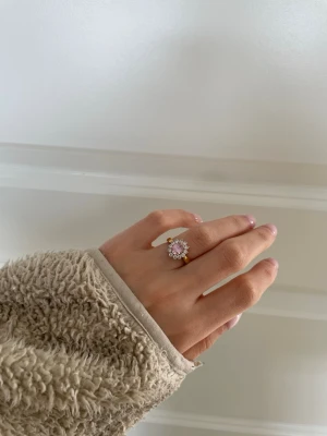 Guldfärgad ring med rosa sten/ guldpläterad - Supersöt guldfärgad ring med en stor rosa sten i mitten, omgiven av små klara stenar i blomformad design. Ringen har en elegant och glittrig look som verkligen sticker ut. Perfekt för dig som vill ha något unikt och färgglatt på fingret.