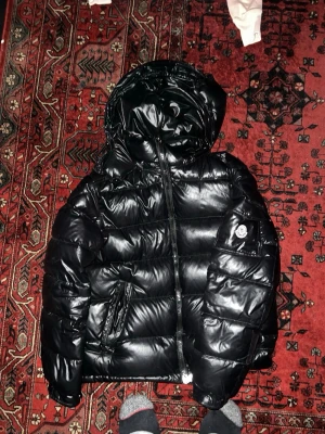 Svart dunjacka från Moncler - Snygg svart dunjacka från Moncler med glansig finish och stor huva.  QR KOD, LABEL INKLUDERAT. Har använts i tvätt och vissa färger i insidan har gått bort. Fickor har en liten skada men fungerar helt bra och bra material 