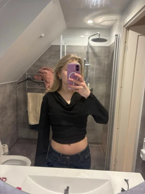 Svart croppad topp från Aware - Säljer en svart croppad topp från Aware med lång ärm och snygg draperad halsringning. Toppen har en tight passform och är tillverkad i mjuk polyester, perfekt till jeans eller kjol. Enkel men trendig design som passar till många olika stilar.