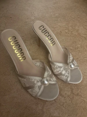 Silvriga sandaler med pärldetalj CUCCOO - Säljer ett par eleganta silvriga sandaler från CUCCOO med öppen tå, smal klack och dekorativ pärldetalj framtill. Ovandelen har ett blommigt spetsmönster som ger en lyxig känsla. Perfekta för dig som vill sticka ut med stilrena detaljer.