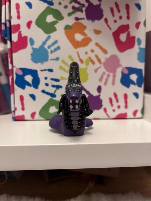 Lila plastfigur orm drake - Säljer en lila plastfigur som föreställer en orm eller drake med svarta detaljer och gula ögon. Figuren har små armar och ett mönster längs ryggen. Perfekt som samlarpryl eller dekoration.
