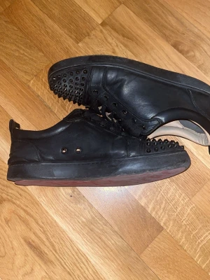 Svarta sneakers med nitar Christian Louboutin - Svarta sneakers i läder från Christian Louboutin med svarta nitar på tån och längs sidorna. Klassisk låg modell med svarta skosnören och röd sula. Insidan är beige och skon har en clean, edgy look.