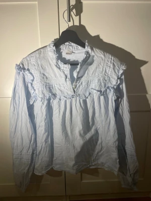 Ljusblå volangblus från Lindex - Söt ljusblå blus från Lindex med volangdetaljer längs axlar och bröst. Blusen har lång ärm, rundad halsringning med liten ståkrage och knäppning framtill. Tillverkad i ett tunt, mjukt bomullstyg som ger en luftig känsla.