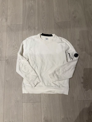 Vit sweatshirt från C.P. Company - En ÄKTA Vit crewneck sweatshirt från C.P. Company med rund hals och ribbade muddar. Tröjan har en ikonisk svart linsdetalj på ena ärmen och en ficka med dragkedja. Perfekt för en clean och stilren look. Tröjan passar även Large. Skriv om ni har några frågor eller om ni vill ha fler bilder