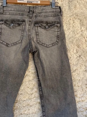 Grå bootcut jeans från gina tricot  - Snygga grå jeans från gina tricot med bootcut passform och slitna detaljer. Jeansen har klassiska fem fickor, dekorativa ficklock bak och silverfärgade knappar. Tillverkade i mjuk bomullsdenim som ger en avslappnad look.