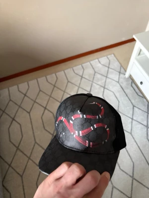 Svart Gucci keps  - Säljer en svart keps från Gucci med det klassiska GG-mönstret och en röd-vit orm på framsidan. Kepsen har böjd skärm och mesh på sidorna för extra ventilation. Perfekt för dig som vill sticka ut med en lyxig accessoar.