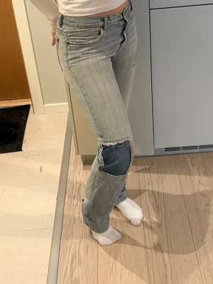 Ljusblå jeans med patchade knän - Säljer ett par ljusblå raka jeans med coola mörkblå patchar över knäna och råa kanter. Jeansen har klassisk femficksdesign och hög midja. Perfekta för dig som gillar en avslappnad och trendig stil. Slitna längst ner då de är väldigt långa i benen