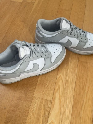 Nike Dunk Low gråvita sneakers - Snygga Nike Dunk Low sneakers i vitt och ljusgrått läder. Klassisk siluett med perforerad tåbox, vita sidopaneler och grå detaljer på snörning, sula och häl. Perfekta för dig som gillar stilrena och tidlösa sneakers.