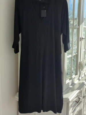 Svart v-ringad klänning från Å+ - Stilren svart klänning från Å+ i storlek L med v-ringning och trekvartsärm. Klänningen har en enkel, rak passform och är perfekt för dig som gillar minimalistisk design. Materialet känns mjukt och lätt mot huden.