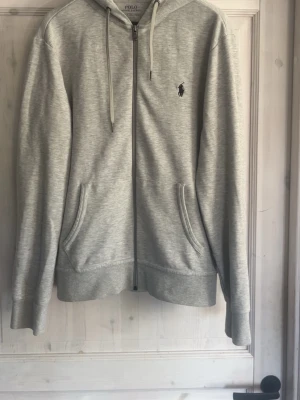 Grå zip hoodie från Polo Ralph Lauren - Snygg ljusgrå hoodie med dragkedja från Polo Ralph Lauren. Klassisk design med huva och dragsnören, två fickor framtill och den ikoniska lilla loggan broderad på bröstet. Tillverkad i mjukt bomullsmaterial som är skönt att ha på sig.