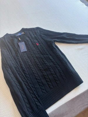 Mörkblå kabelstickad tröja Polo Ralph Lauren - Kabelstickad mörkblå tröja från Polo Ralph Lauren med rund halsringning och ribbade muddar. Klassisk röd broderad logga på bröstet. Tillverkad i mjukt material som passar perfekt till lager på lager. Snygg och tidlös design. 