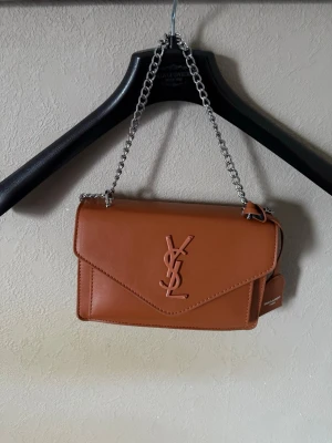Brun YSL handväska i läder - Snygg brun handväska från YSL i slätt läder med silverfärgad kedjerem. Väskan har ett klassiskt kuvertformat lock och den ikoniska YSL-loggan framtill. Perfekt storlek för det viktigaste och ger en lyxig touch till din outfit.
