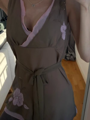 Brun omlottopp med blommor och knyt - Säljer en brun omlottopp med ljusrosa blommor och matchande knytband i midjan. Toppen har v-ringning med rosa kant och är ärmlös, perfekt för en trendig och feminin look.