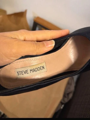 Svarta pumps med strass från Steve Madden - Snygga svarta pumps från Steve Madden med glittriga strassdetaljer på klacken. Skorna har en klassisk form och är tillverkade i mocka för en lyxig känsla. Perfekta för dig som vill sticka ut med stilrena men ändå glamorösa skor.